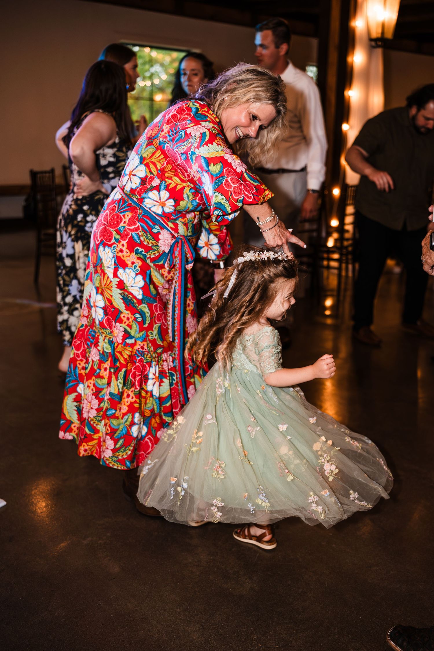 Woman in colorful floral dress dancing with young flower girl in sage green tulle dress at wedding.