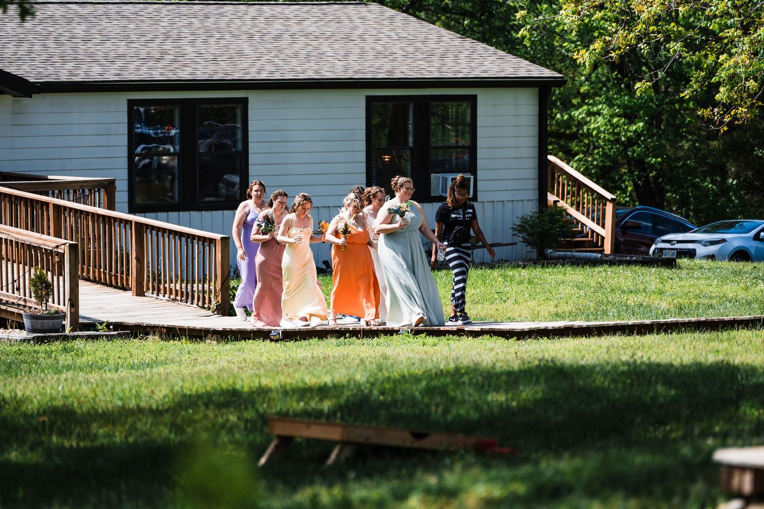 Bridesmaids in colorful mismatched gowns walk together across a grassy lawn toward a white farmhouse venue.