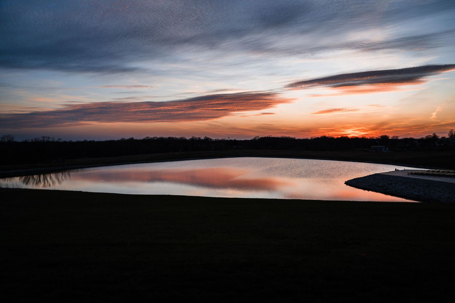 Serene sunset reflecting on a calm lake with vibrant orange and pink hues illuminating the dramatic cloudy sky.