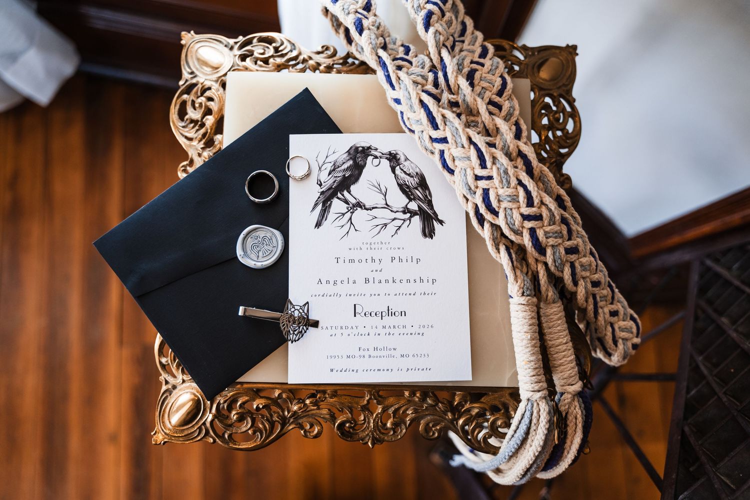 Elegant wedding invitation with bird illustration, wax seal, rings, and braided rope on ornate gold tray.