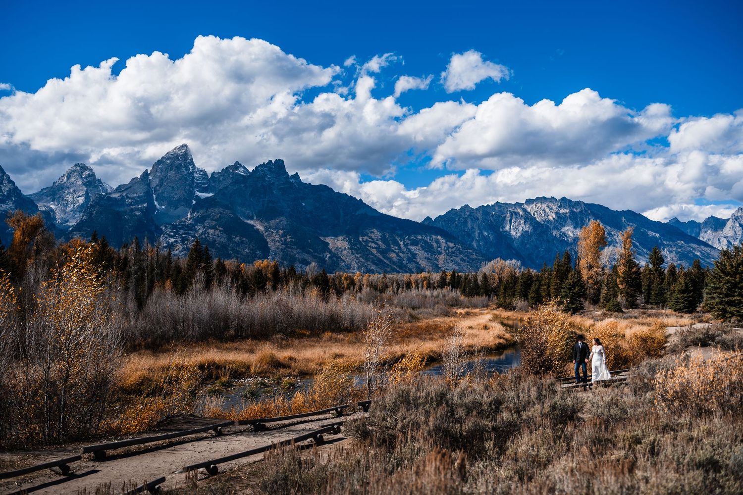 Panoramic view of the Teton mountain range with golden autumn fields and dramatic clouds against a blue sky.