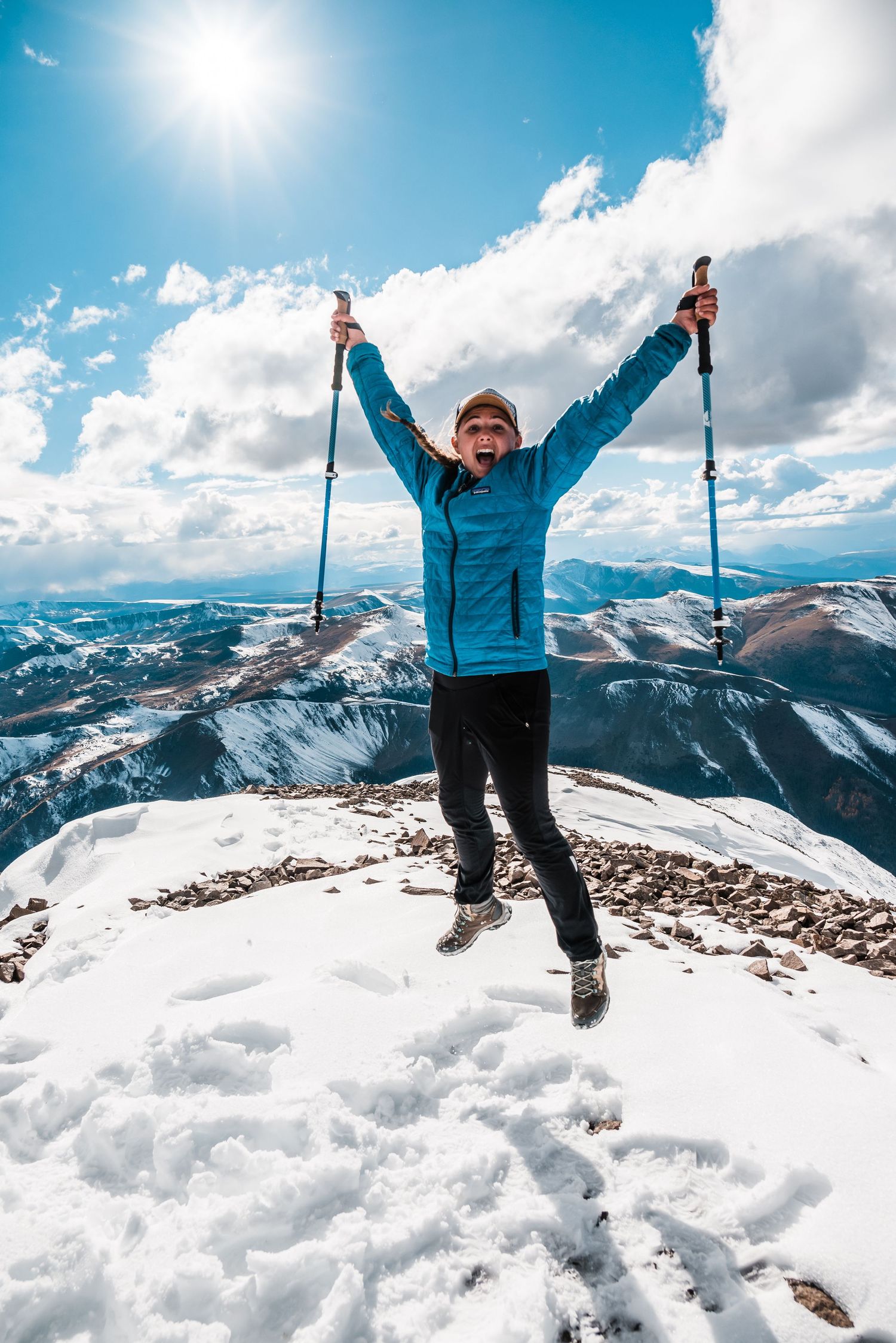 Person celebrating on snowy mountain peak against bright blue sky and snow-capped peaks.