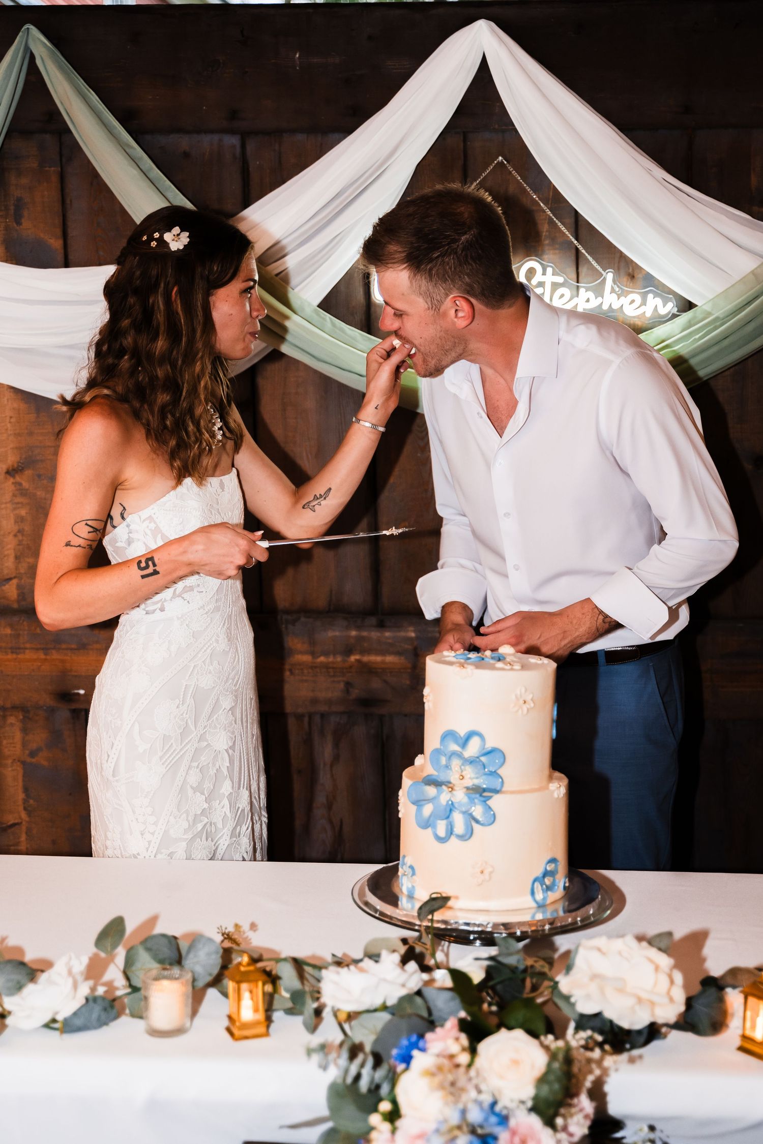 A couple shares a playful moment while cutting their two-tier wedding cake decorated with blue floral details.