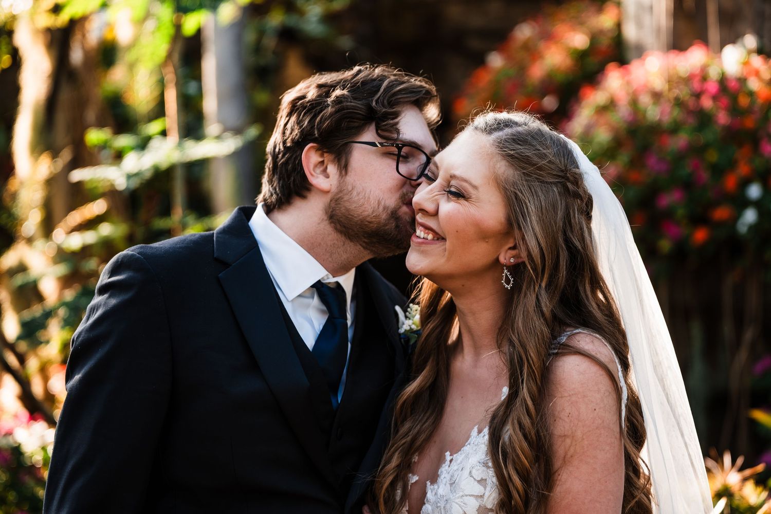 Wedding couple laughs together during outdoor portrait session with pink flowers in background.