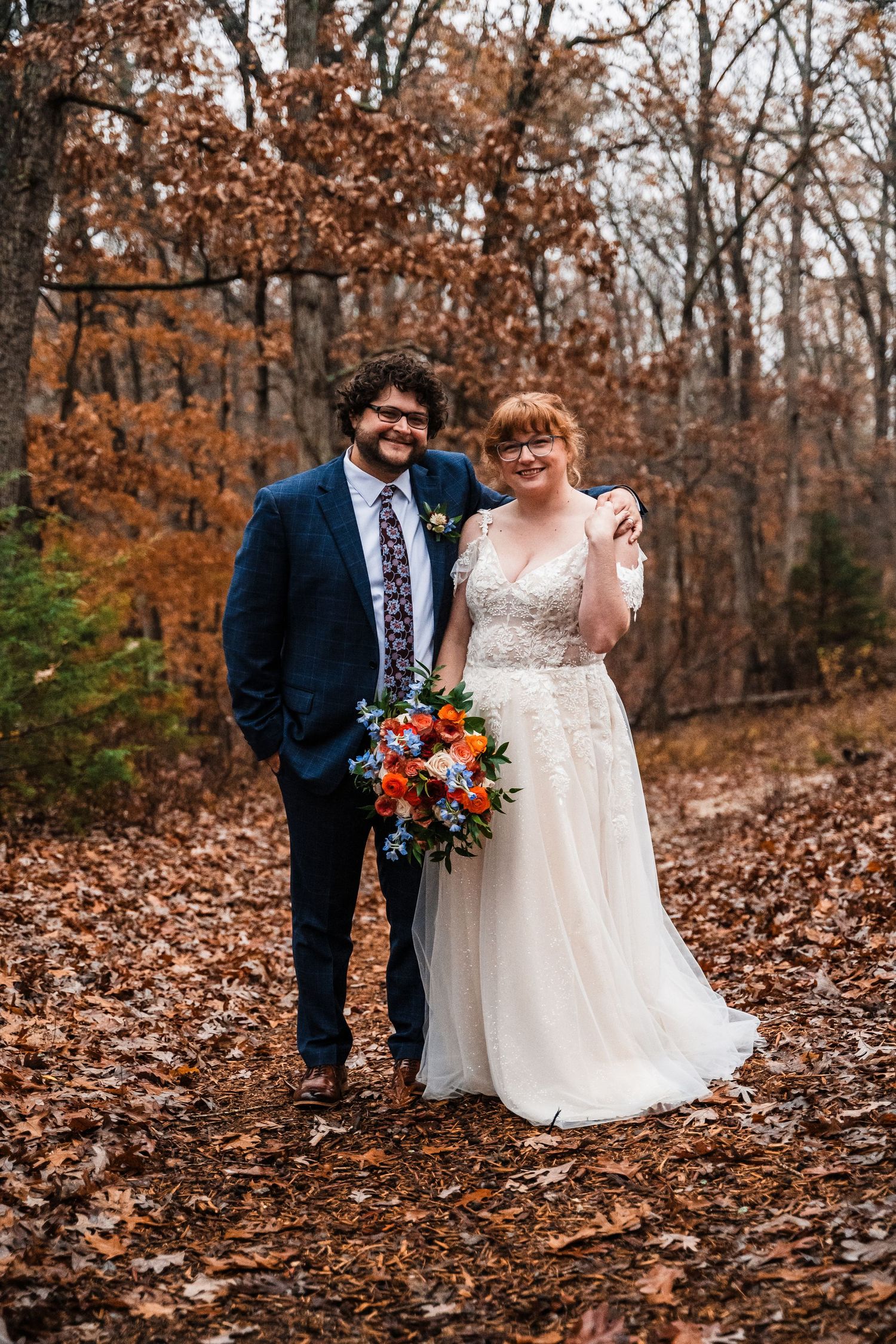 Autumn wedding portrait with couple standing among fallen leaves in wooded setting.
