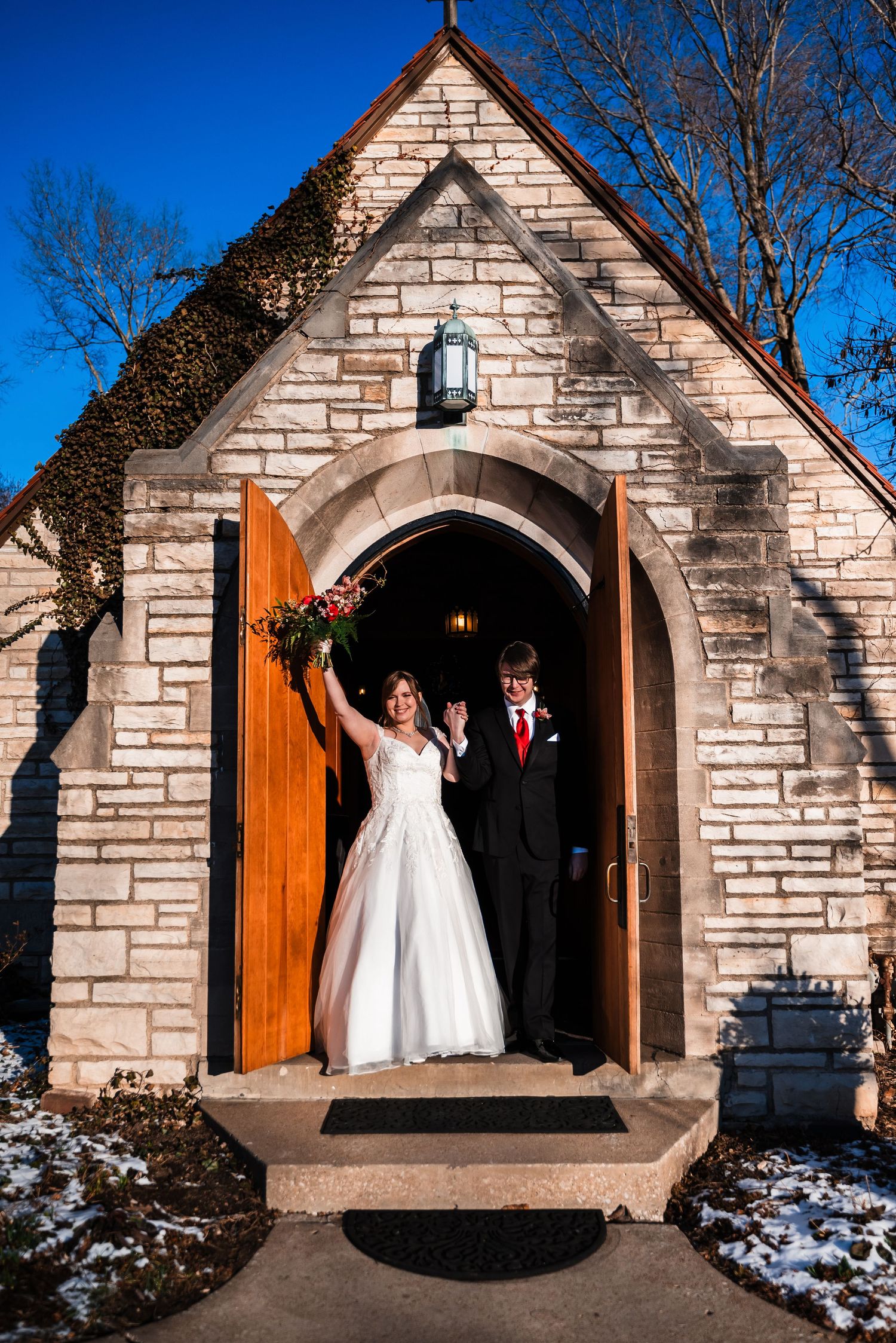 Newlyweds celebrate joyfully in the doorway of a small stone chapel with snow on the ground.