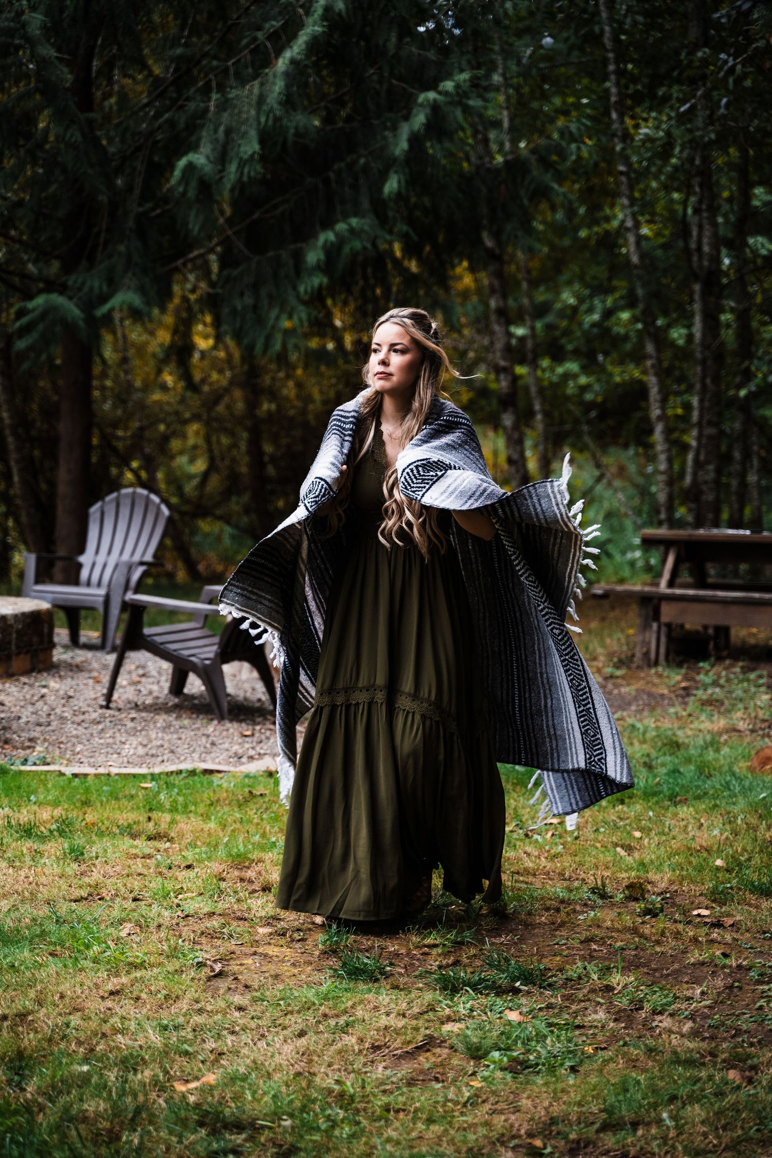 A long-flowing olive dress and grey cardigan create a whimsical forest scene with Adirondack chairs in the background.