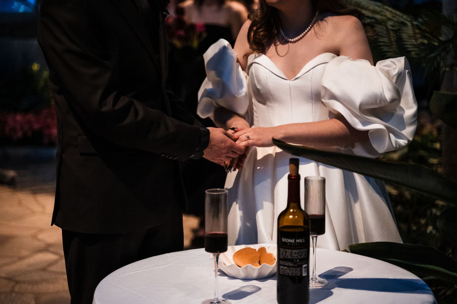 Close-up of hands during a wedding ceremony with wine and appetizers on a white table.