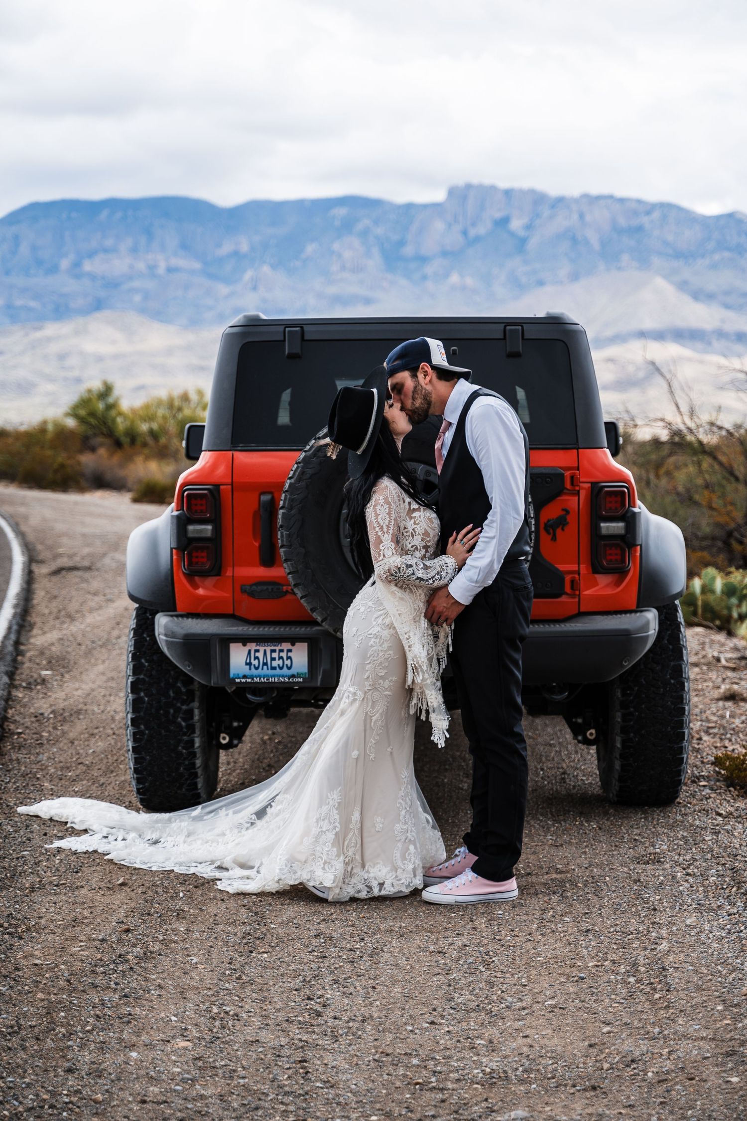 A wedding couple poses beside a red Jeep Wrangler in a desert landscape with mountains in the background.