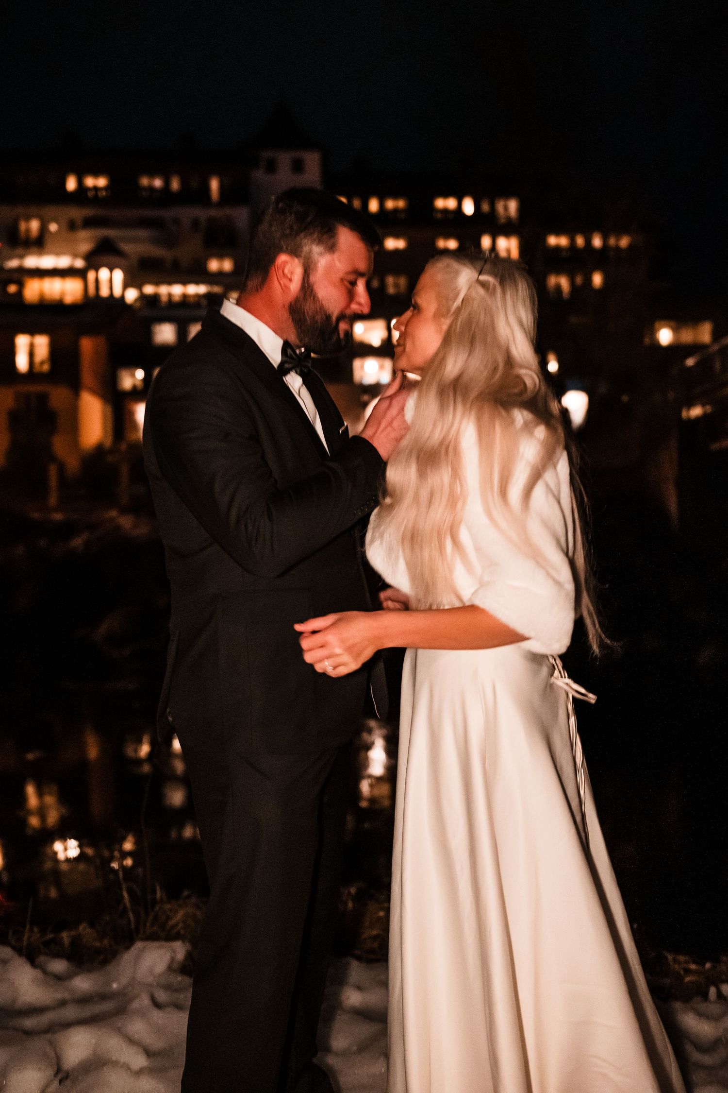 A romantic nighttime wedding portrait with warm lights from buildings glowing in the background.