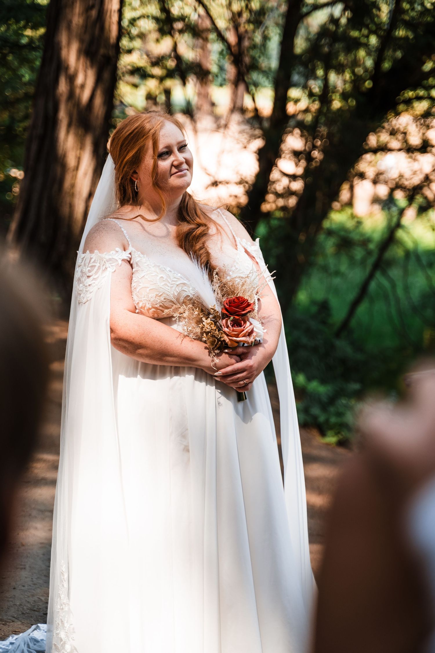 A bride in a flowing white cape dress holds a bouquet while standing in a sunlit forest setting.