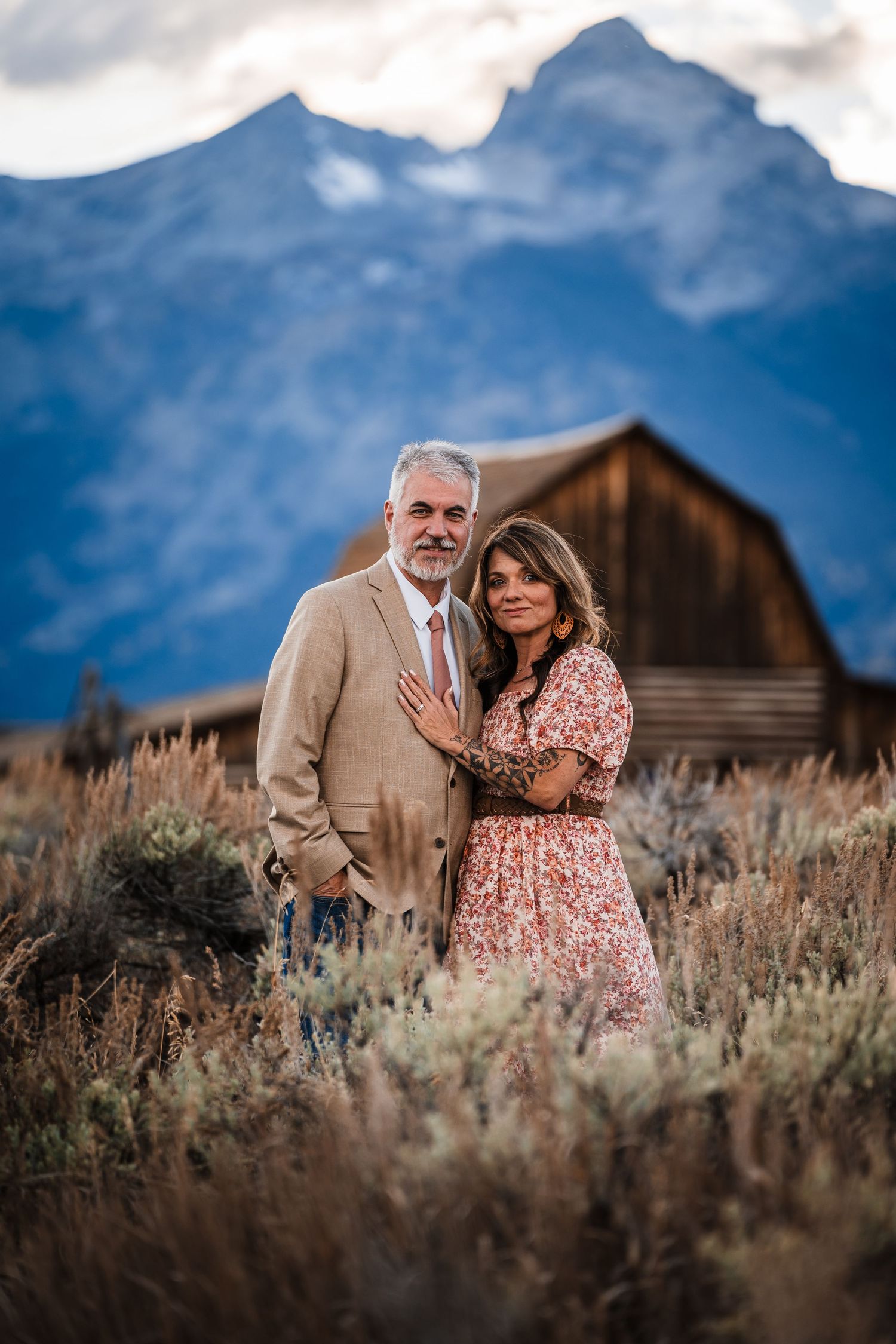 A couple embraces in a field of sagebrush with rustic wooden buildings and mountains in the background.