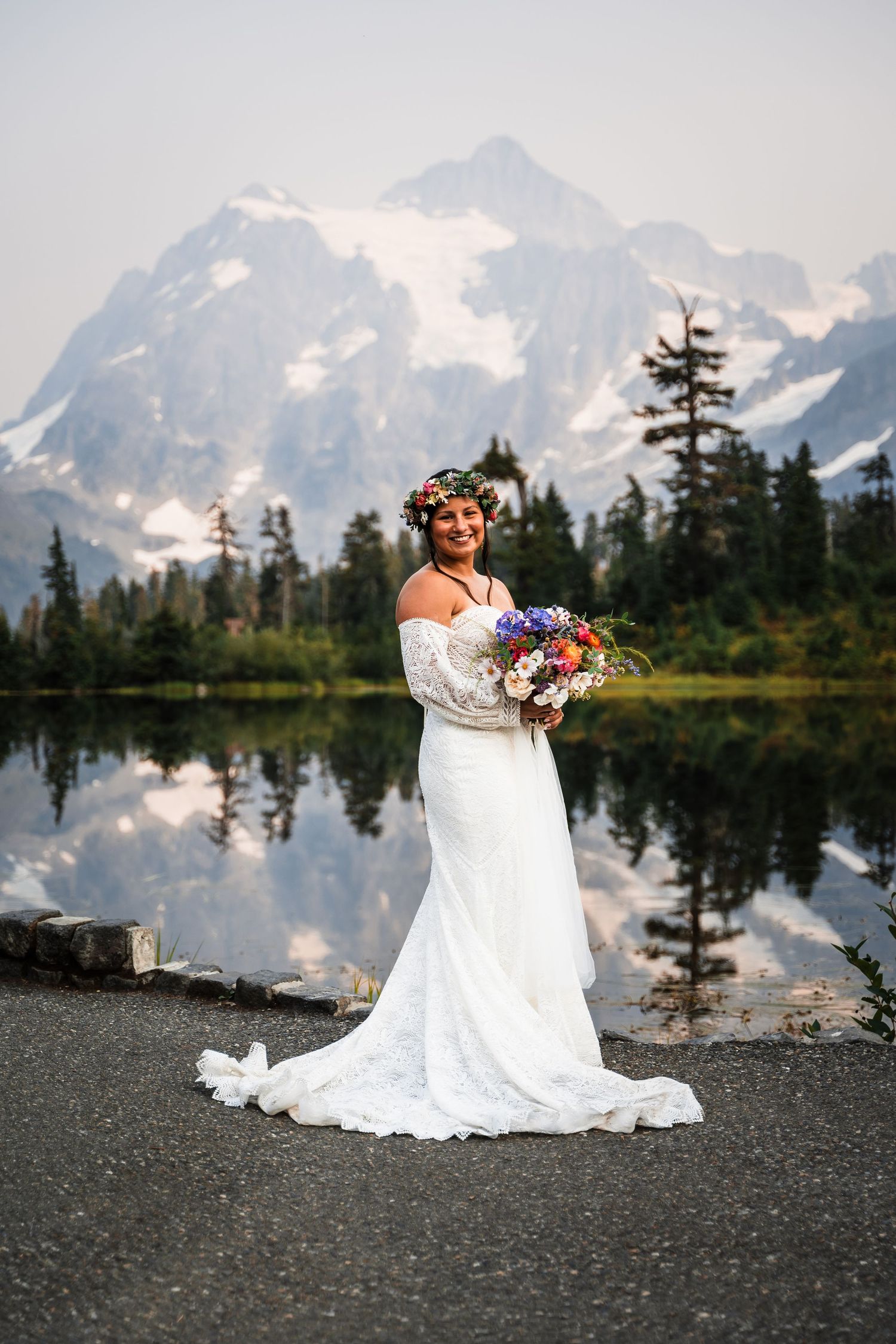 A white wedding dress flows against a stunning mountain lake backdrop with snow-capped peaks and evergreen trees.