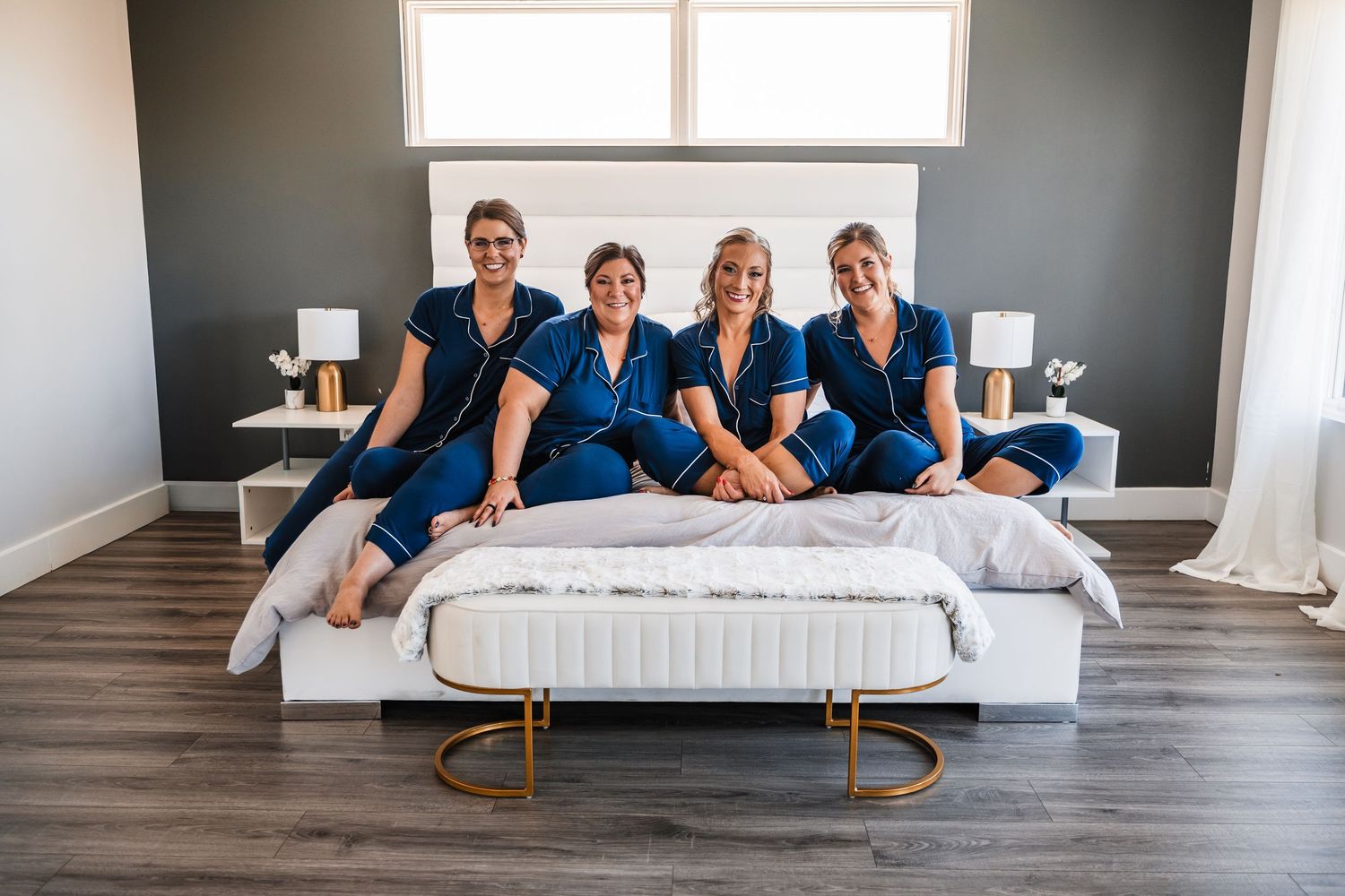 Four people wearing matching navy blue outfits sit together on a white bed in a modern bedroom with grey walls.