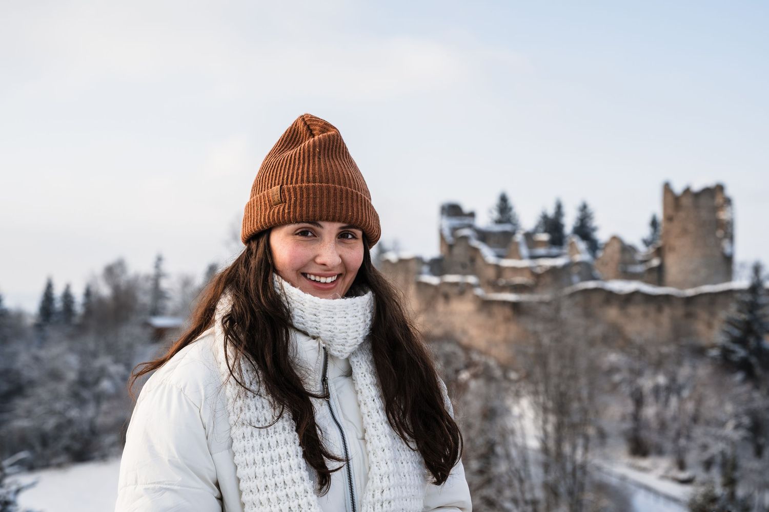 Person wearing a white knit sweater and brown beanie smiles while enjoying a winter mountain landscape.