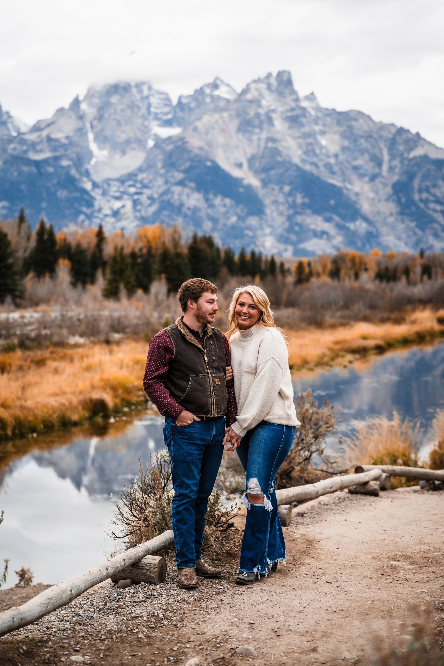 A couple enjoys a scenic autumn view along a river trail with the Grand Teton mountains rising majestically in the background.