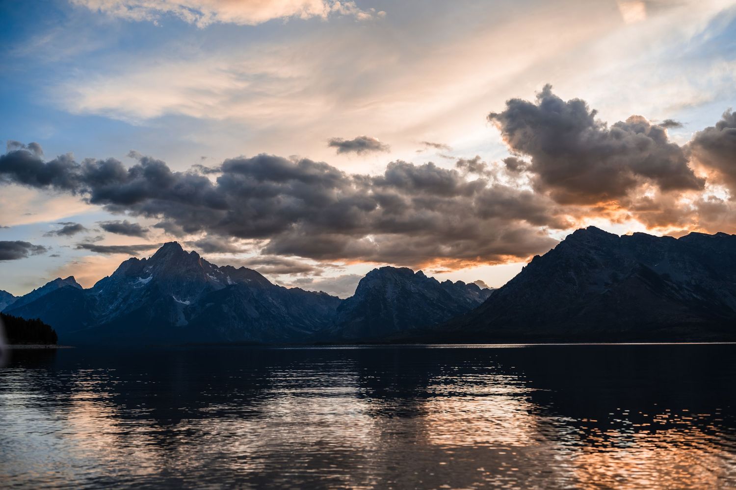 Mountain silhouettes reflect on calm water during dramatic sunset.