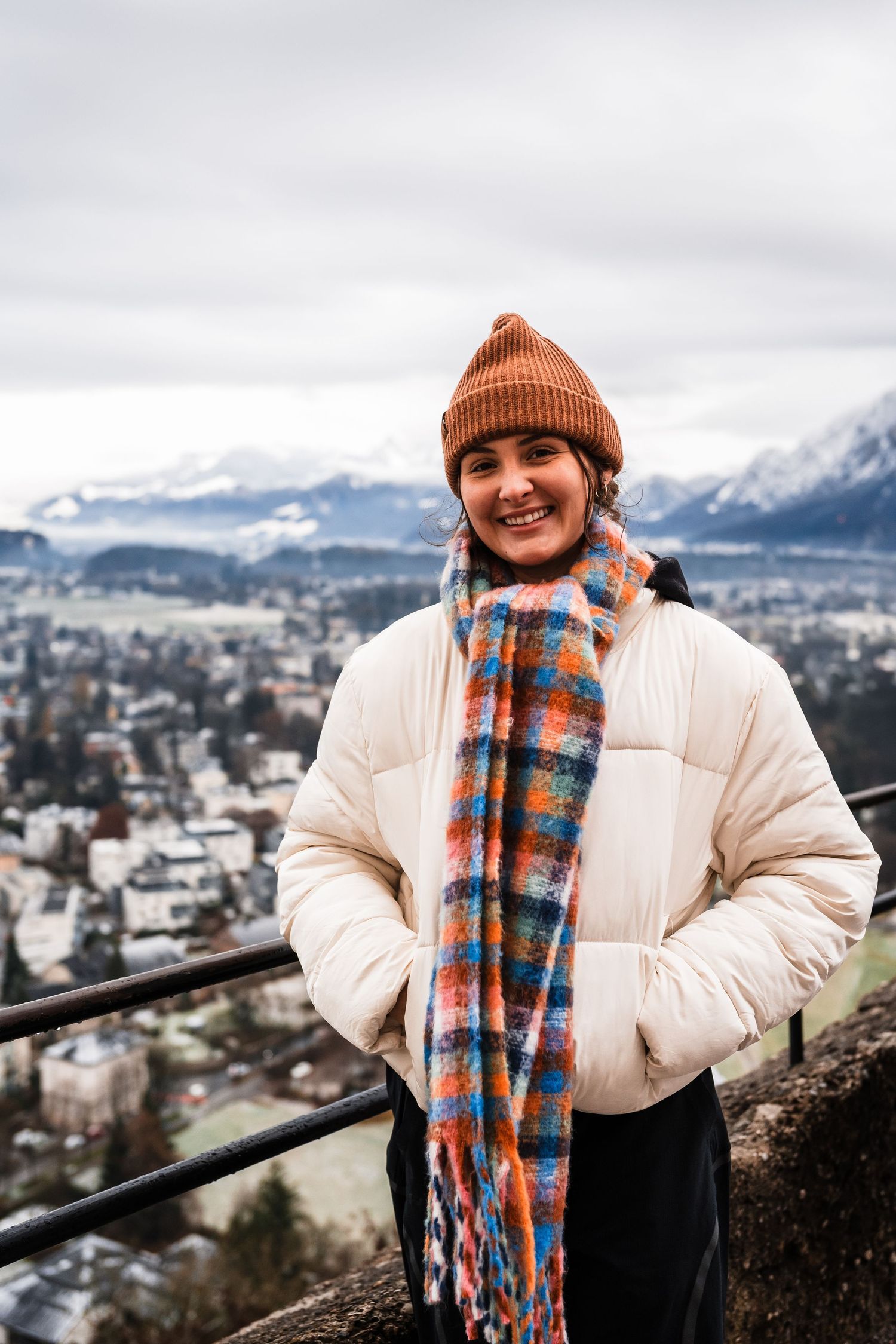 Person in a cream puffer jacket and orange beanie poses against a snowy mountain backdrop.