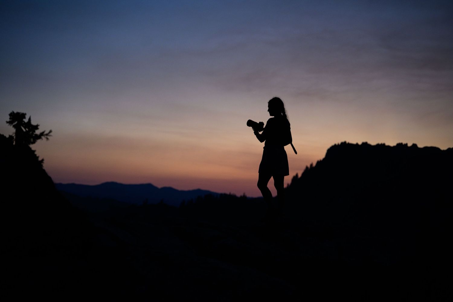 Silhouette of a woman holding a camera on a rocky hilltop at sunset with colorful sky and mountain range backdrop.