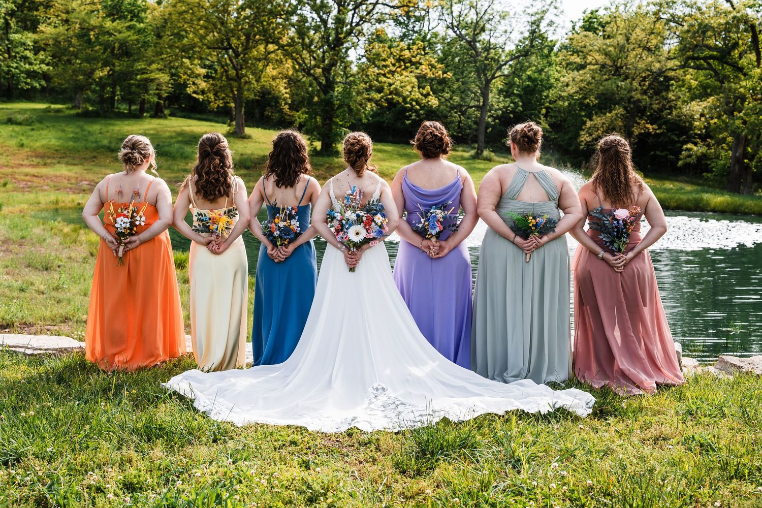 Bride with colorful bridesmaids in mismatched dresses pose with bouquets by a pond.