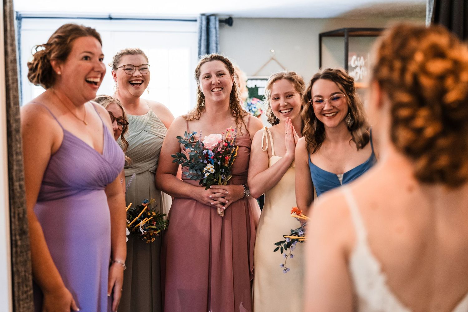 Bridesmaids in mixed purple dresses laugh seeing bride in mirror for first time.