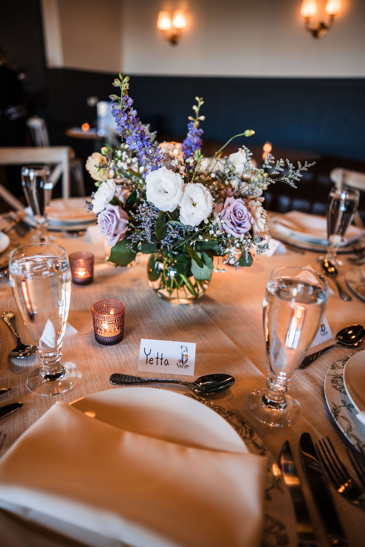 Elegant wedding reception table with purple and white floral centerpiece, place cards, and candlelight ambiance.