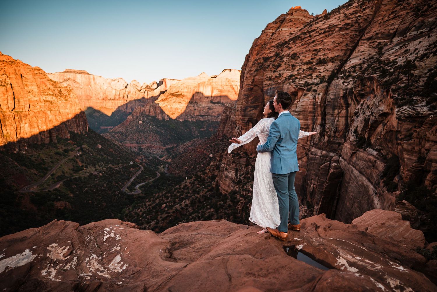 Bride and groom standing on cliff edge at Zion National Park overlooking stunning canyon at sunset.