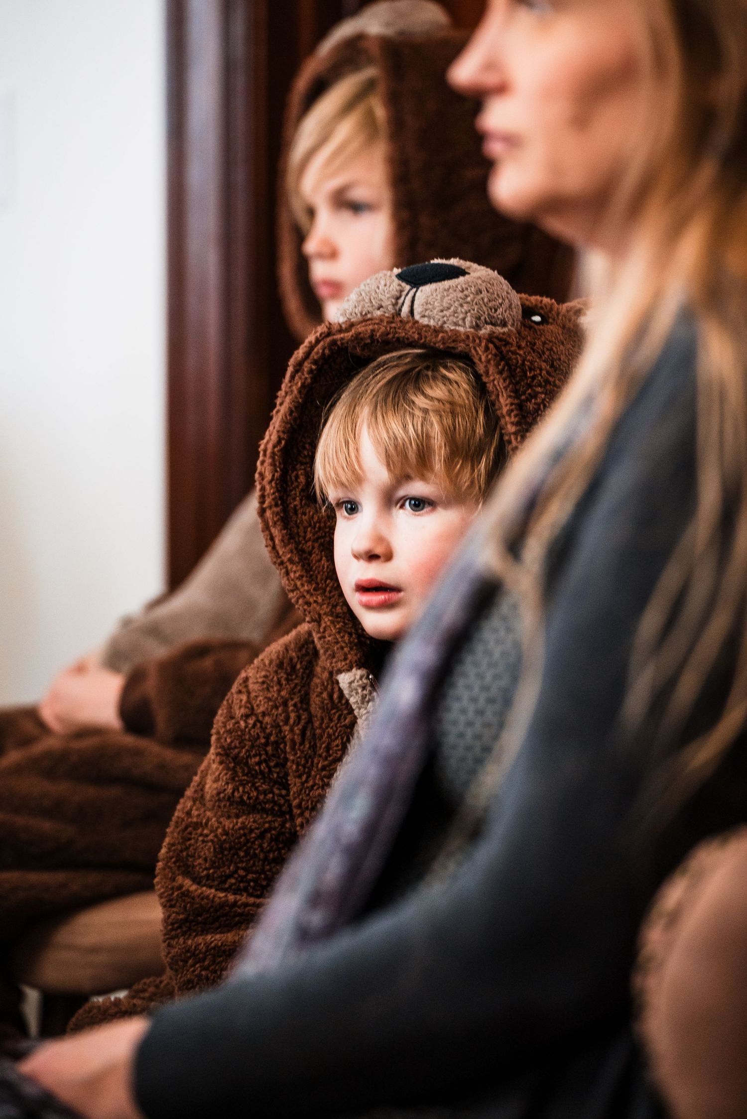 Young child in brown bear costume hoodie sits between two blonde women in a warmly lit indoor setting.