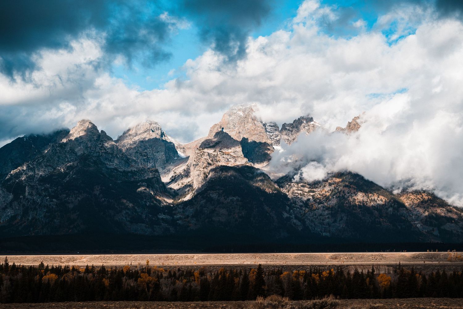 Dramatic mountain landscape panorama with snow-capped peaks under moody blue and white clouds stretching across horizon.