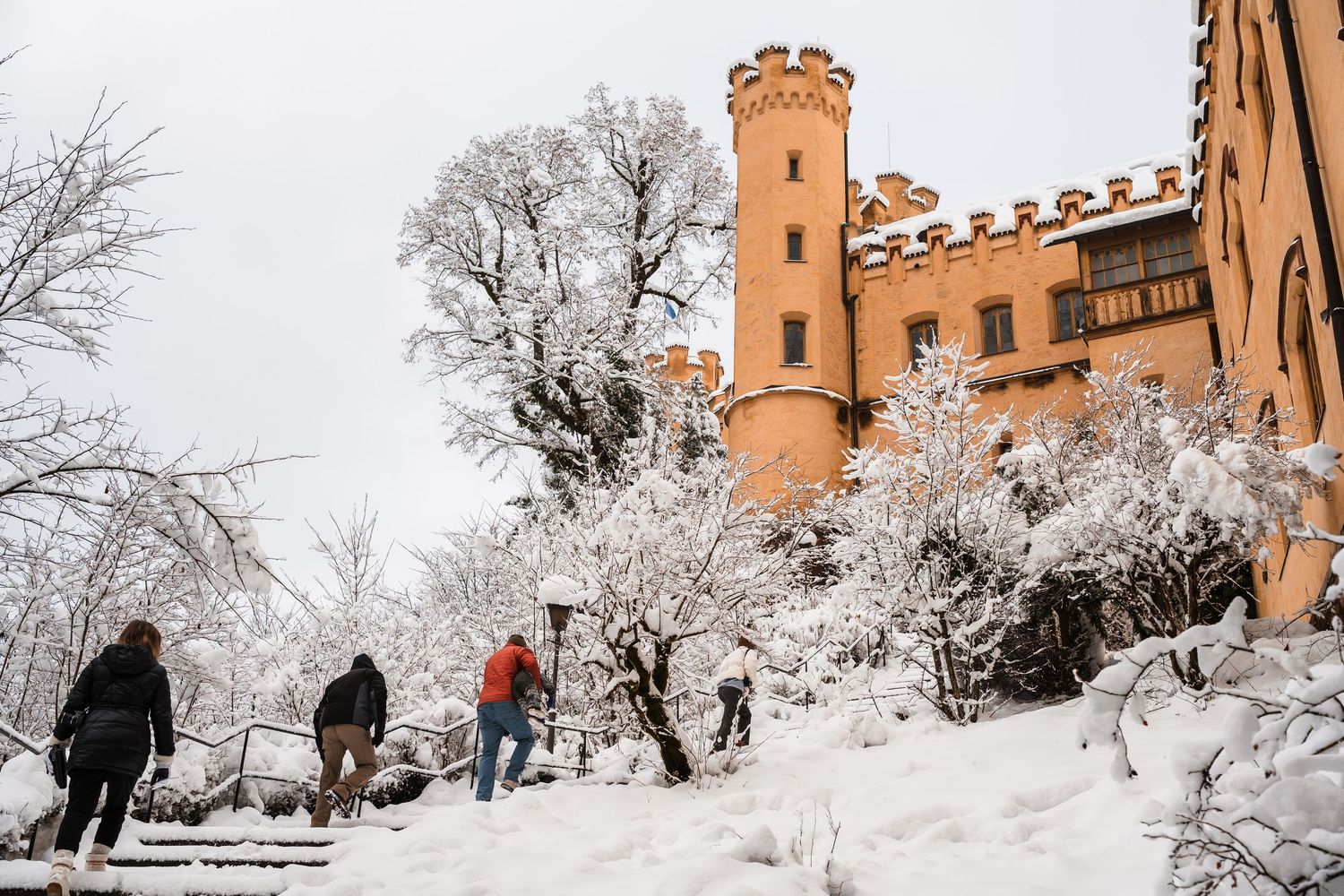 People enjoy sledding down a snowy hill near an orange brick castle-like building on a winter day.