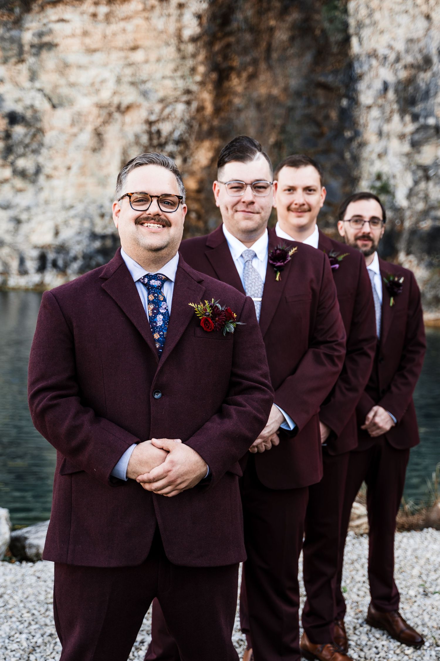 Groomsmen in burgundy suits pose together by a stone wall for wedding photos.