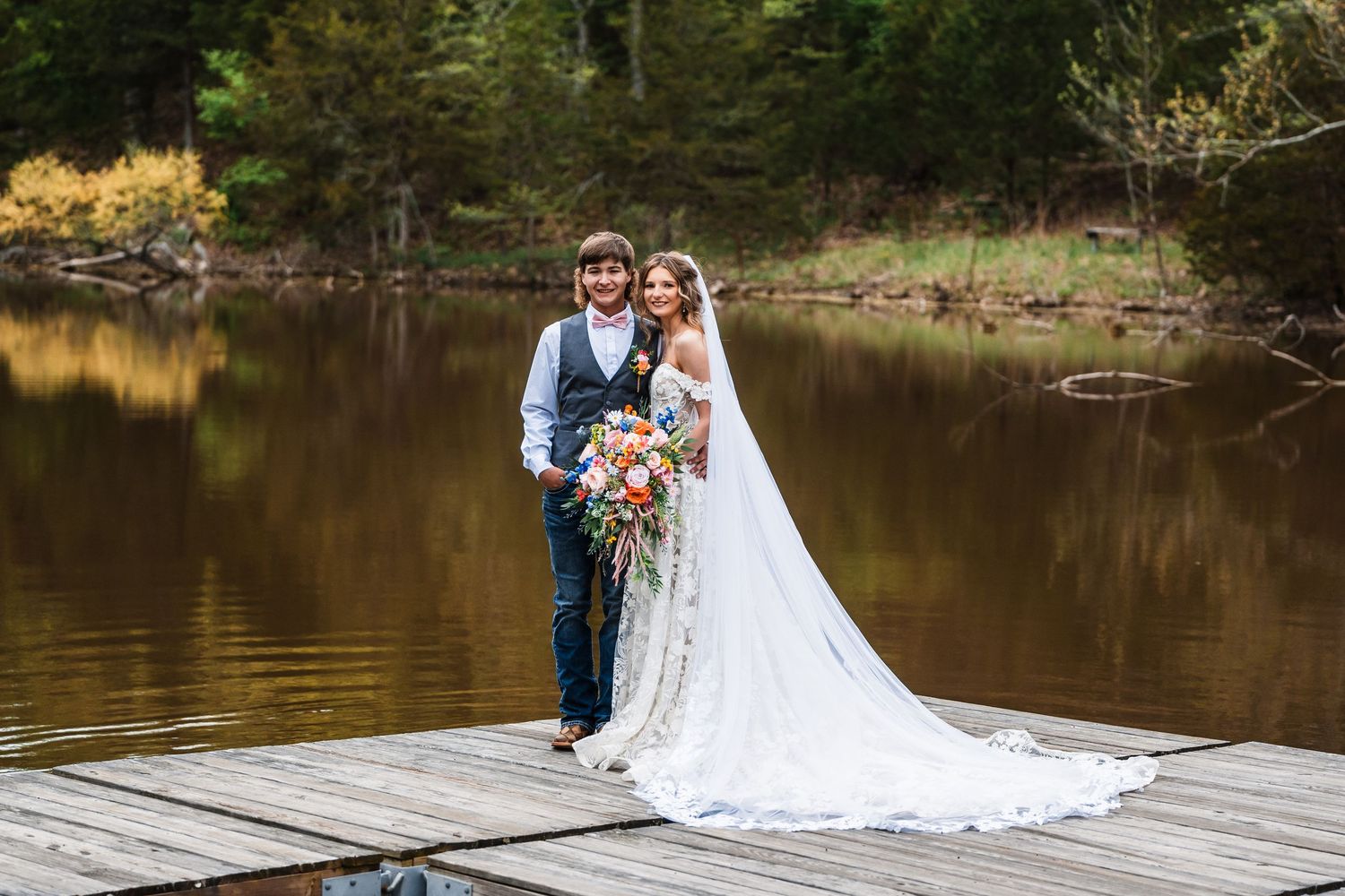 A couple poses on a wooden dock by a serene lake, with a flowing white wedding dress trailing behind.