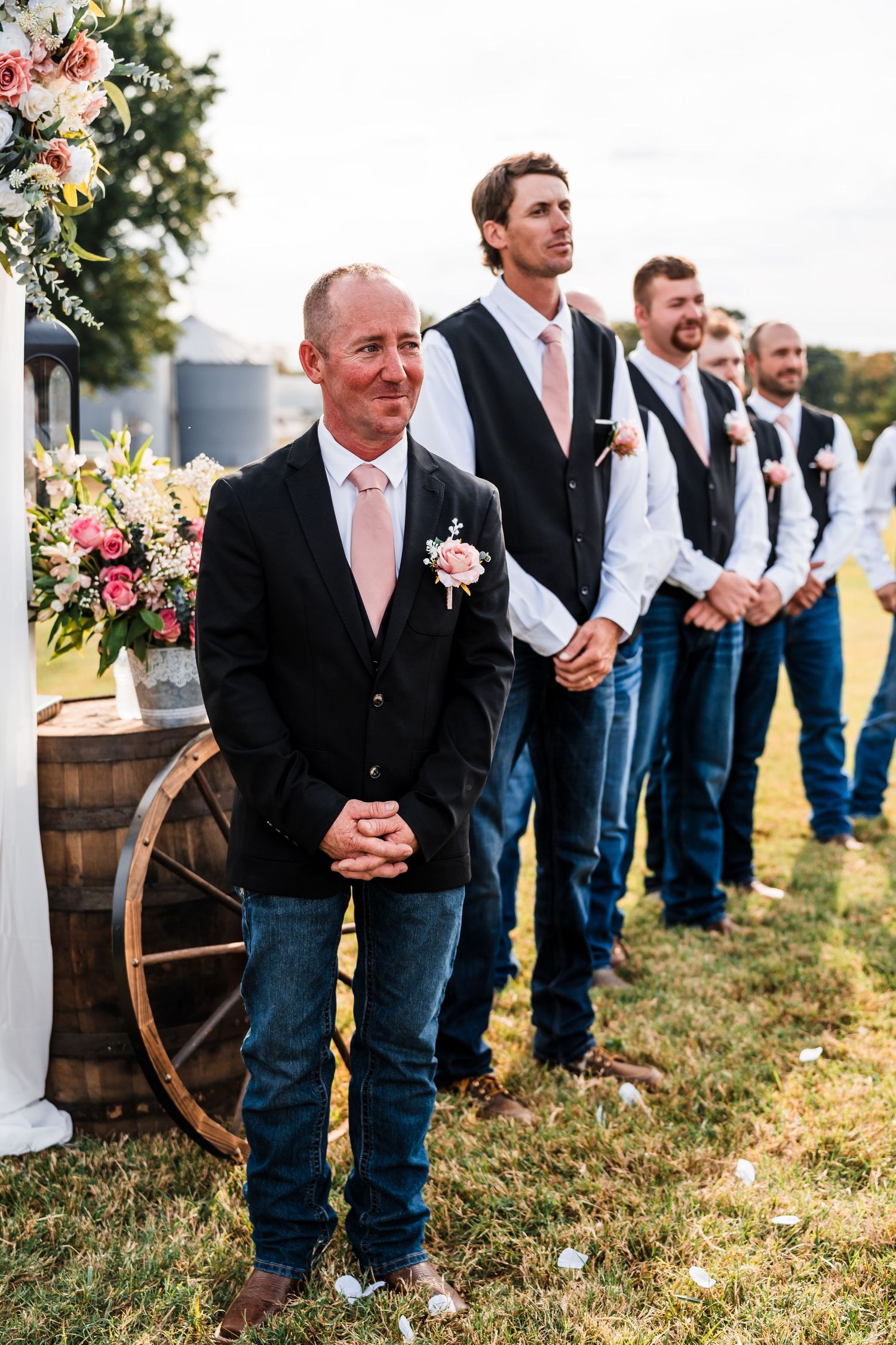Groomsmen in casual attire with jeans and vests lined up outdoors at rustic wedding venue.