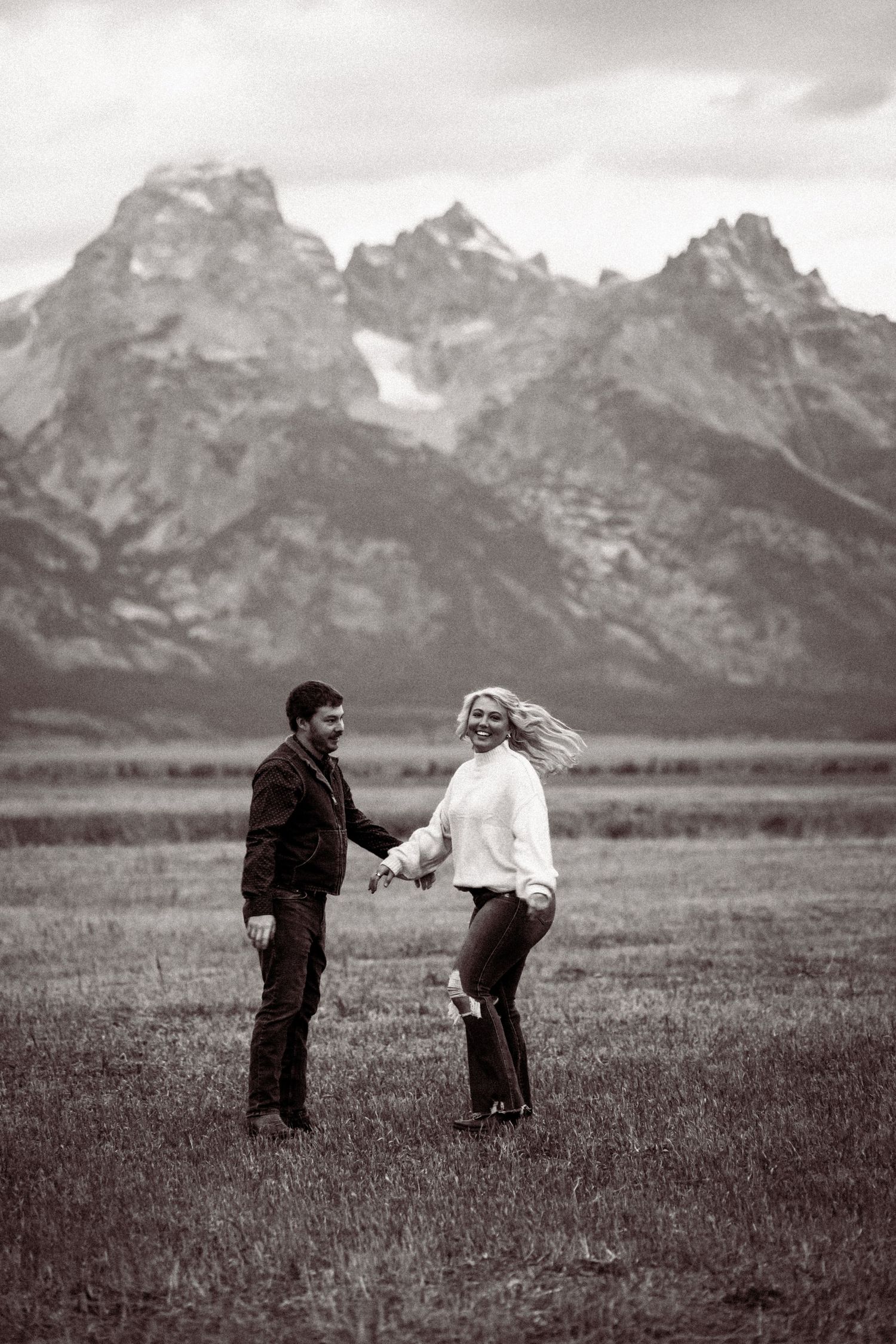 Black and white photo of two people playfully interacting in a field with the Grand Tetons towering in the distance.