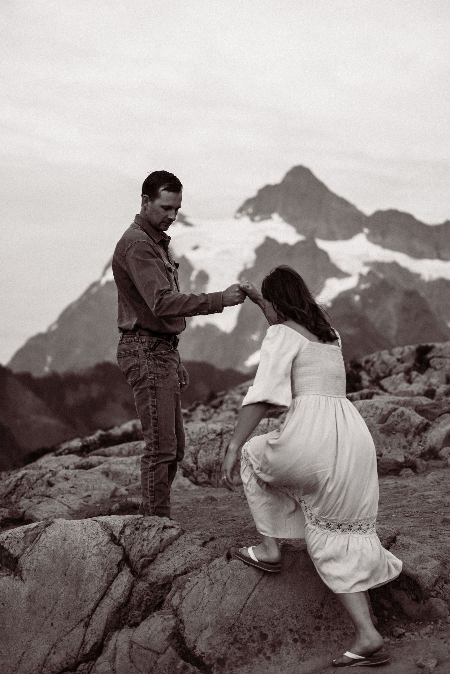A black and white photo captures a romantic moment between a couple on rocky terrain with a majestic mountain backdrop.
