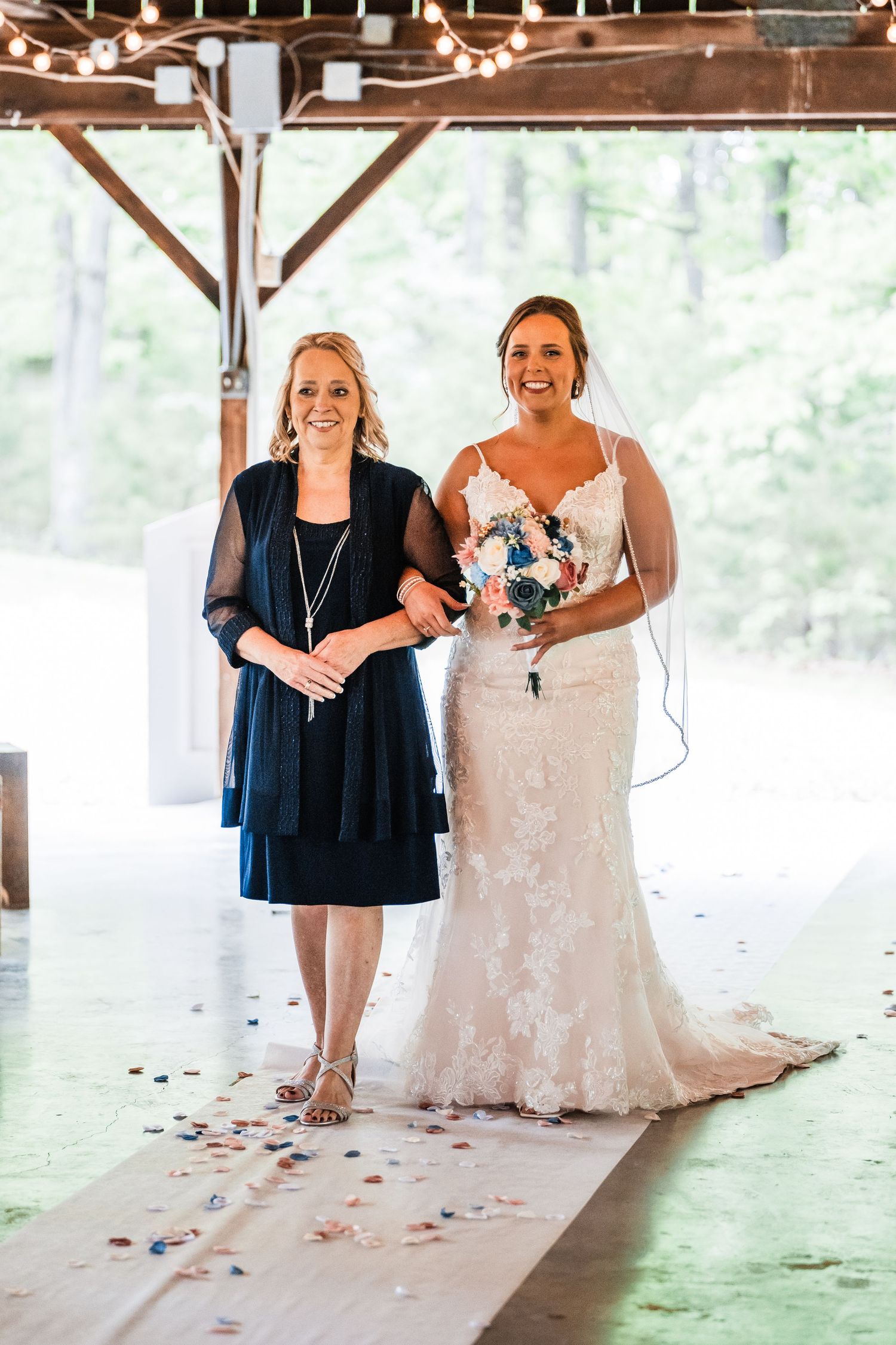 Two people stand under a covered venue walkway decorated with rose petals, one wearing a navy dress and one in a white gown.