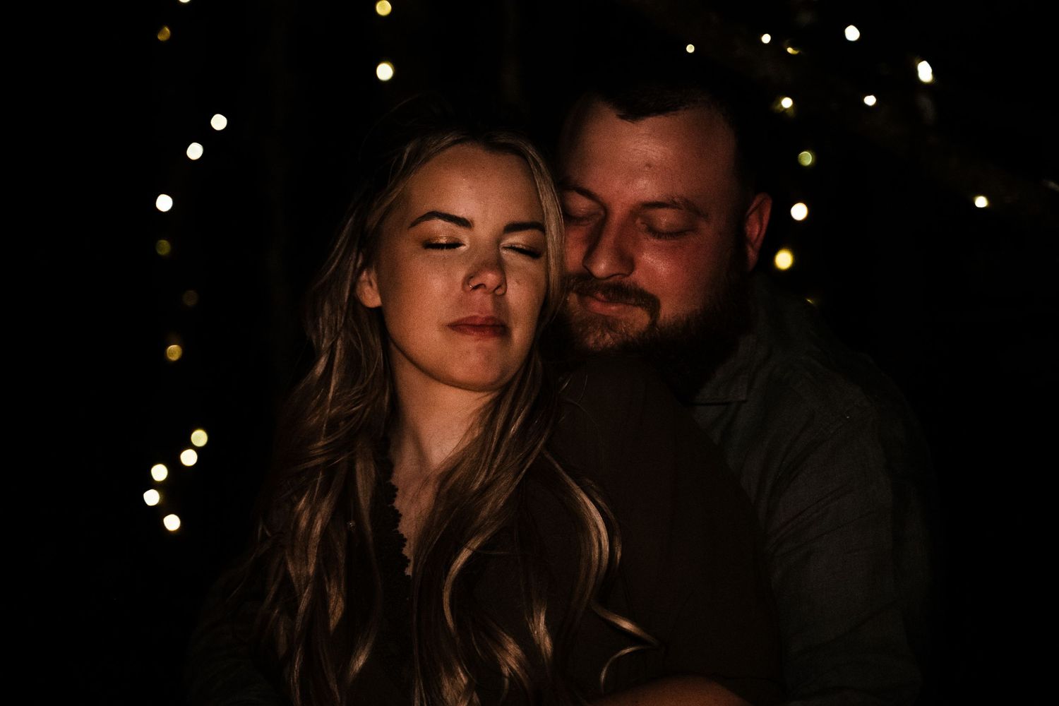 Intimate romantic couple embraces in the dark with twinkling bokeh lights in the background.