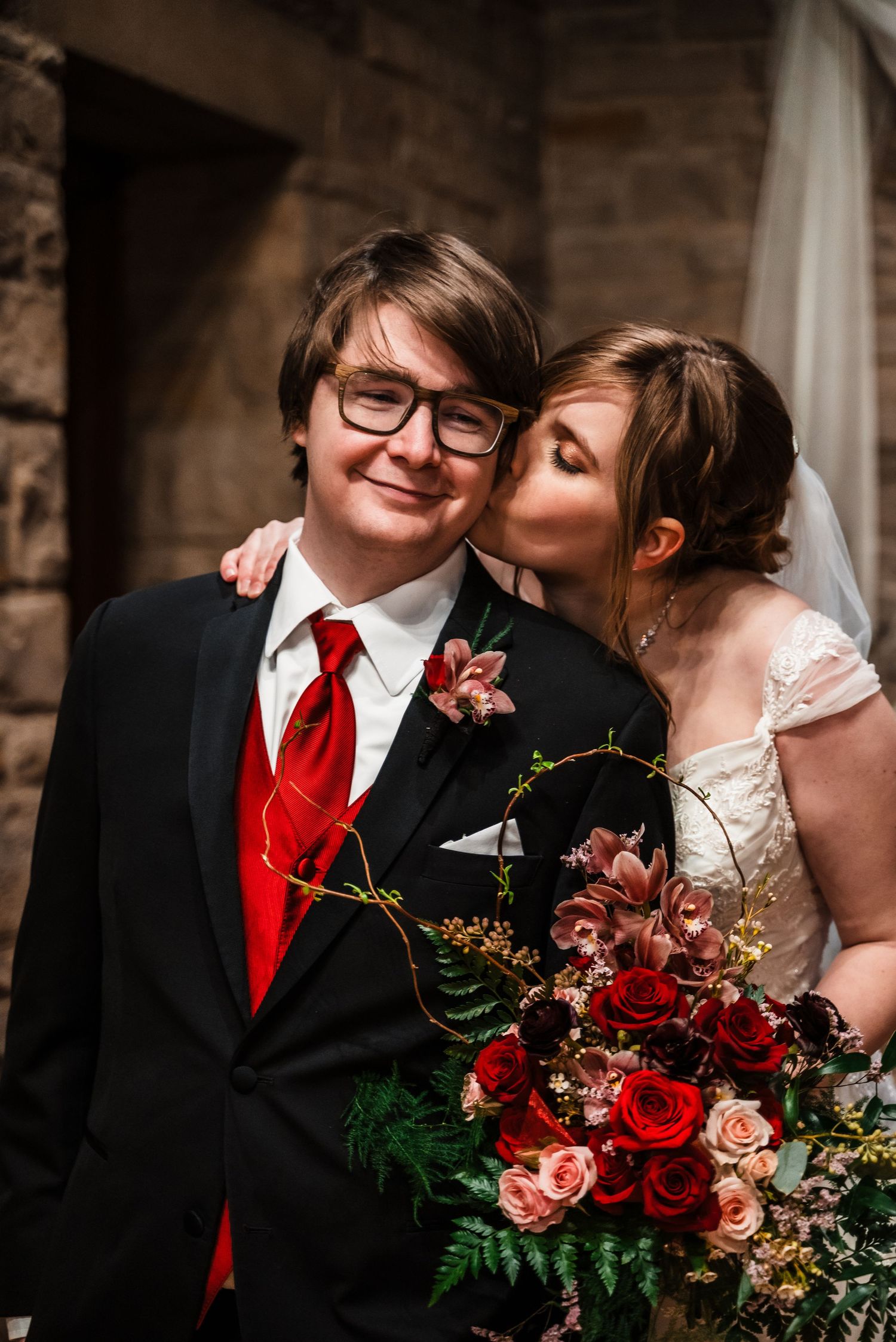 A wedding couple with red roses and a vibrant bouquet pose together in a stone chapel.