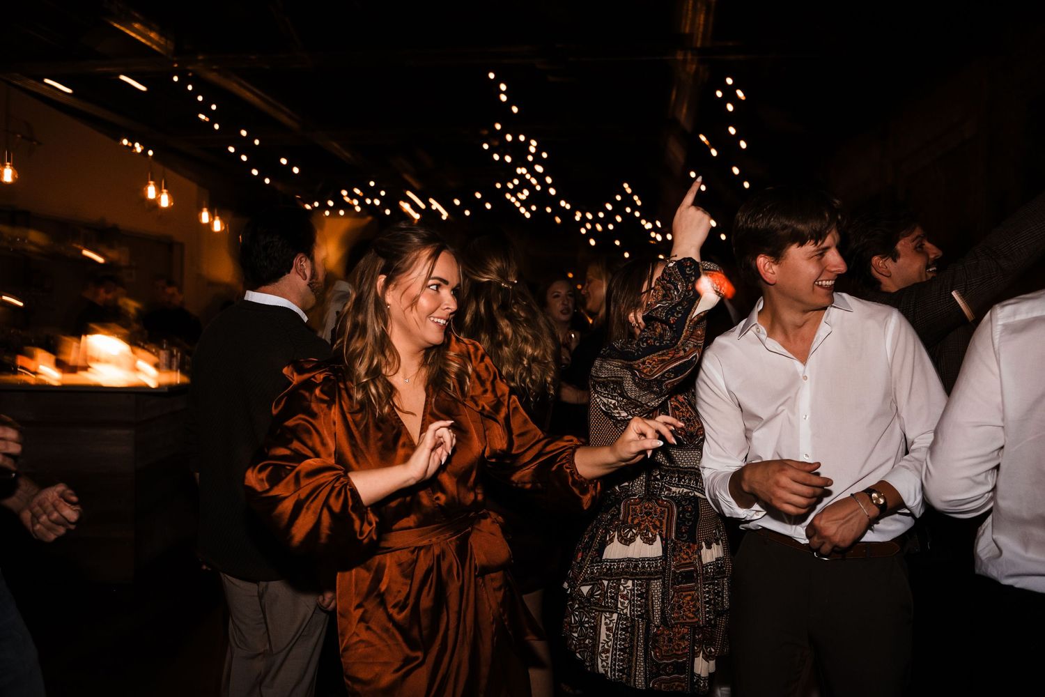 Partygoers dancing and celebrating under decorative string lights at an indoor venue.