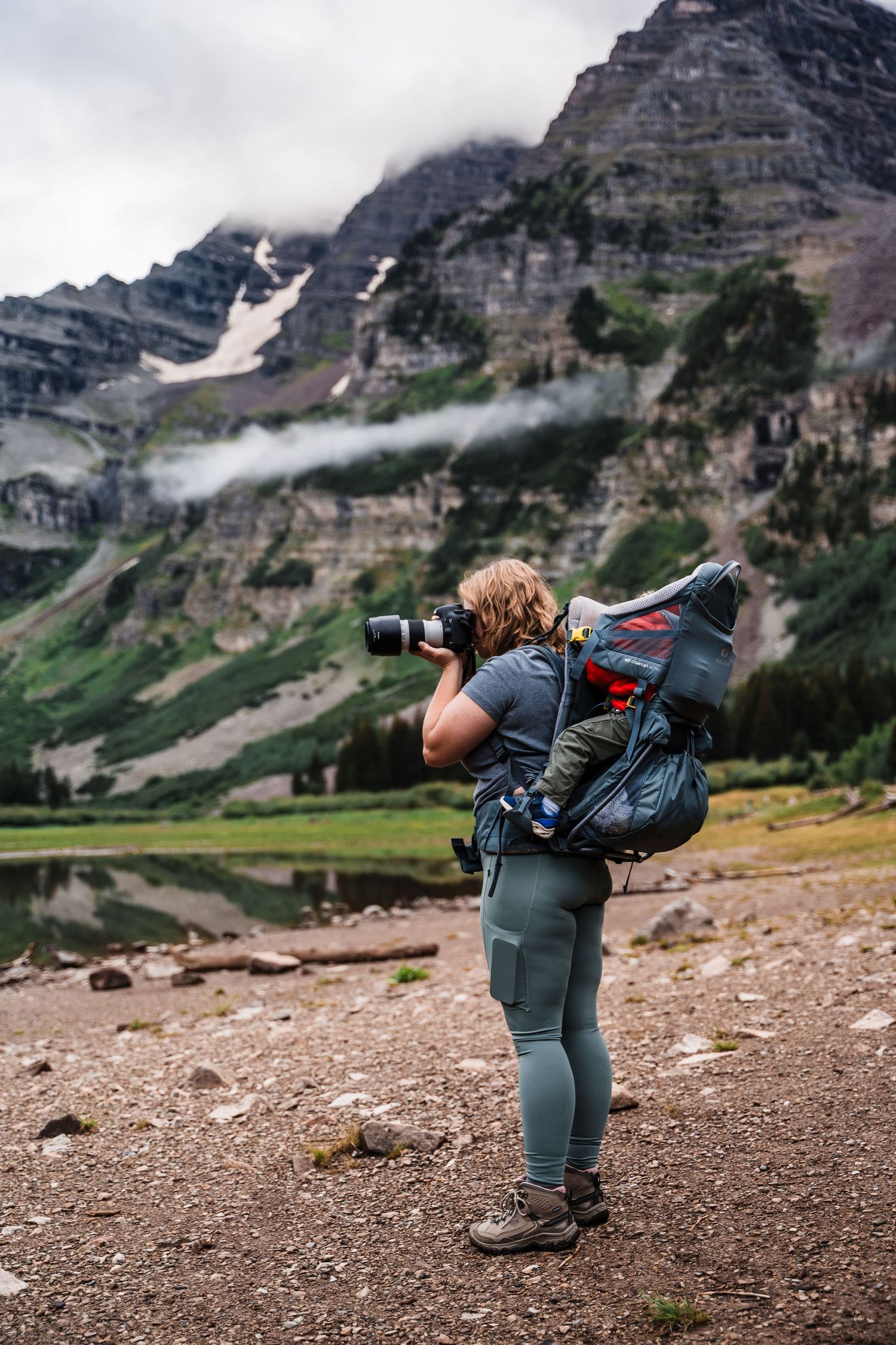 Photographer with gear captures mountain scenery beneath dramatic rocky cliffs and misty clouds.