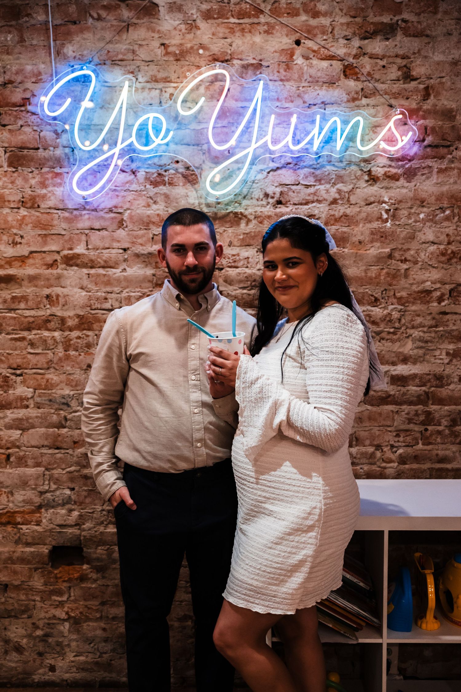 A couple poses beneath a blue neon 'Yo Yums' sign against an exposed brick wall.