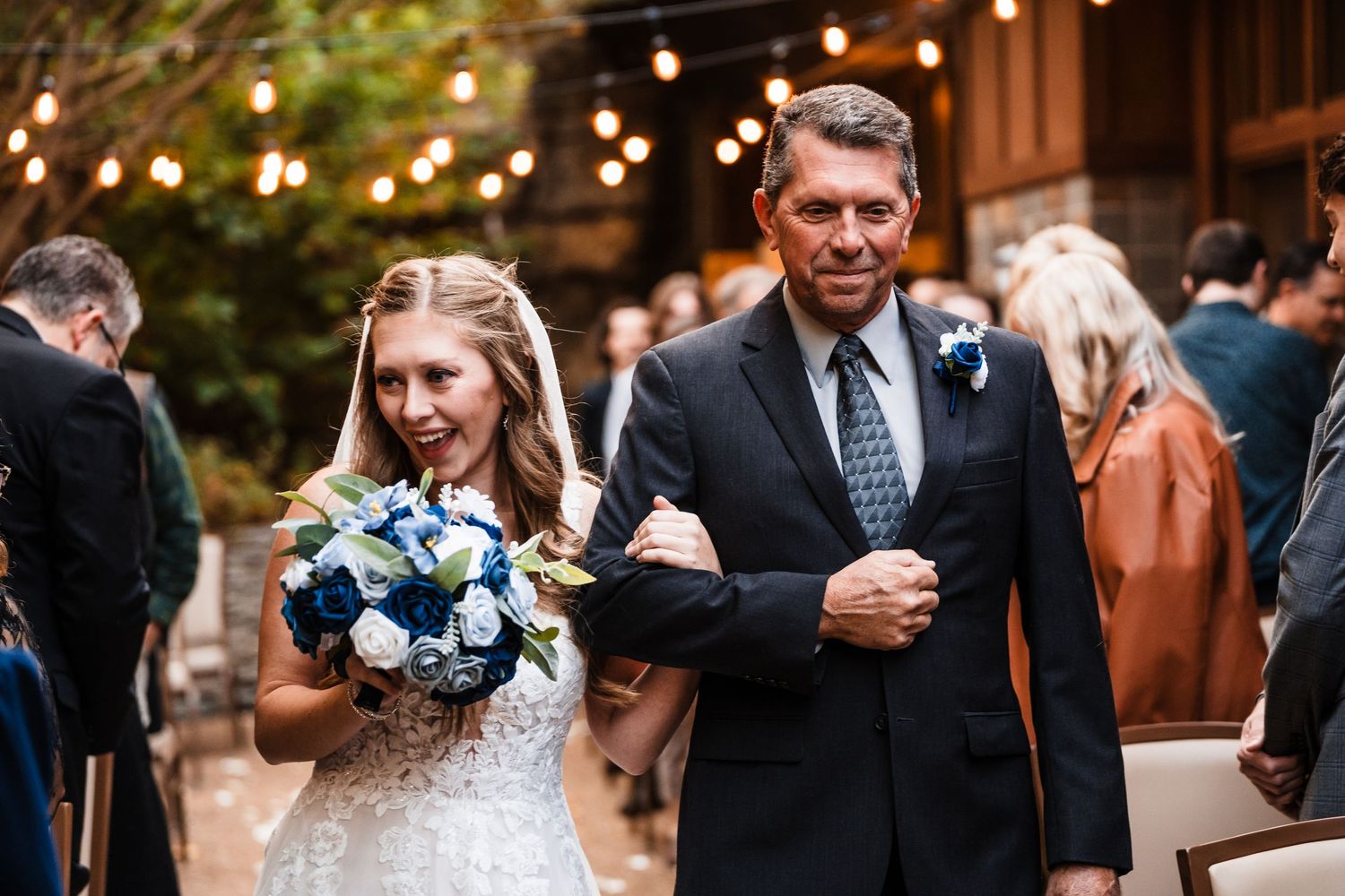 Wedding processional along petal-lined aisle under romantic string lights at outdoor venue.