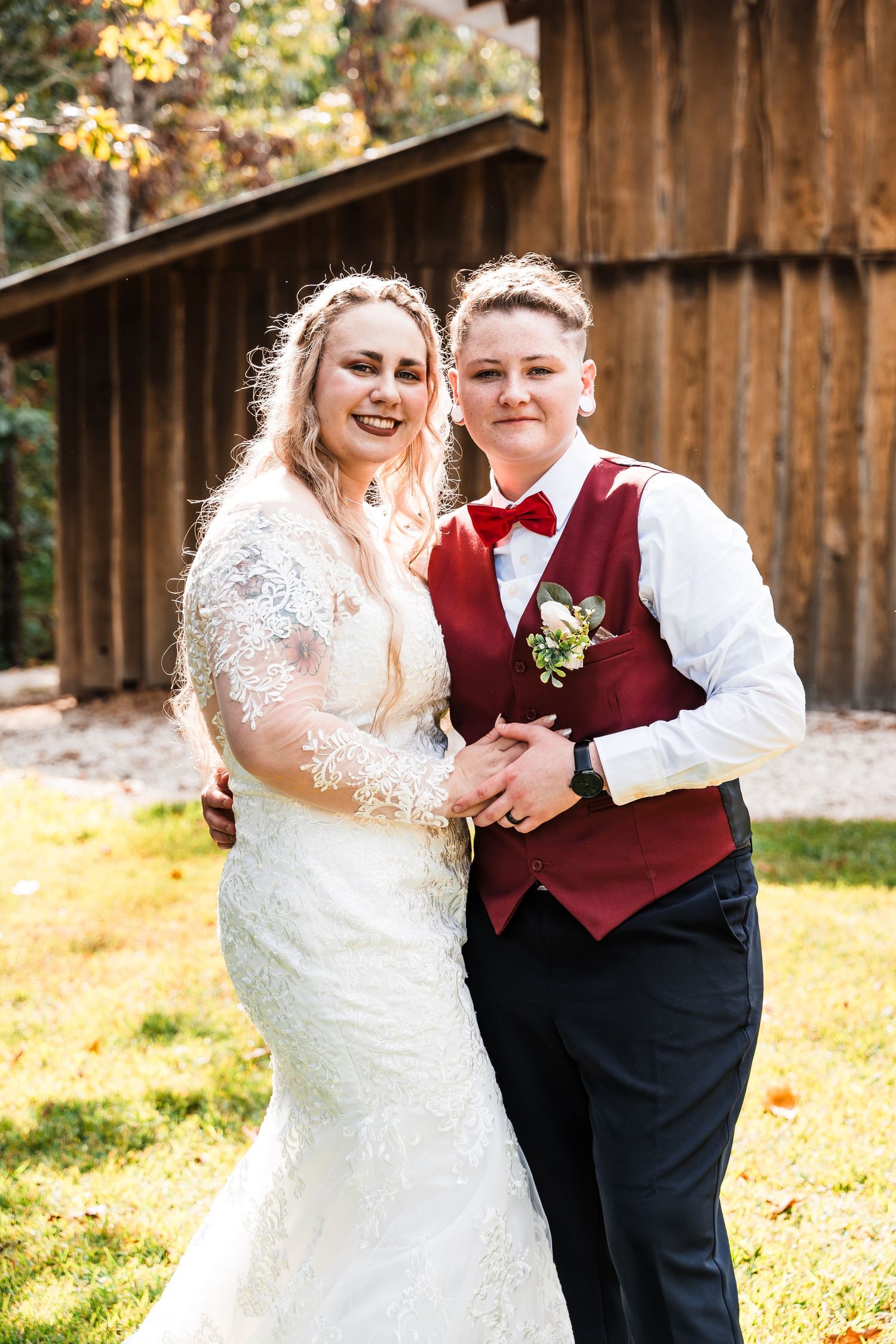 Newlywed couple in formal attire smiling outdoors with rustic barn backdrop.