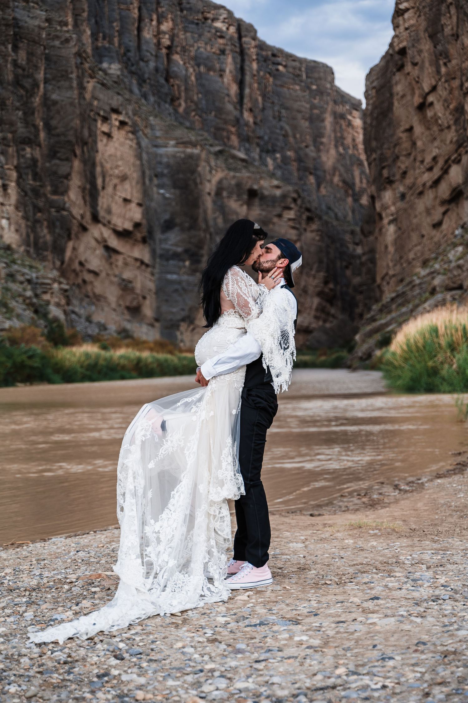 A couple embraces by a river with towering canyon walls creating a dramatic backdrop.