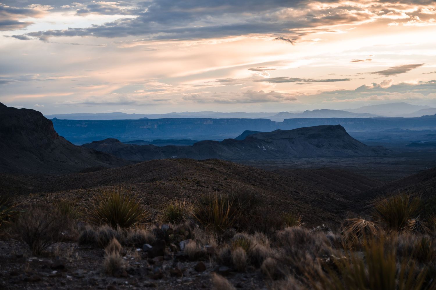Dramatic sunset over rugged mountain landscape with dark silhouettes and desert vegetation in foreground.