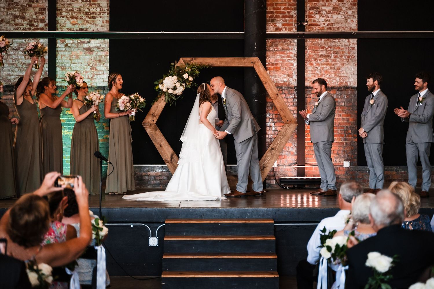 Wedding ceremony takes place in a rustic brick venue as couple shares a kiss beneath an arch.