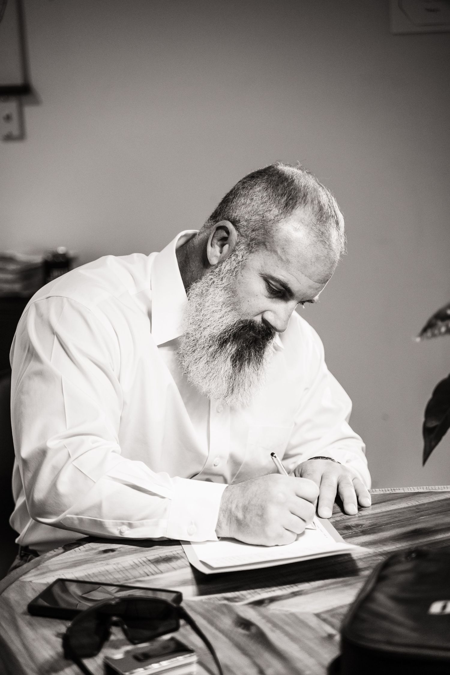 A bearded man in white shirt writes in a notebook at a desk.