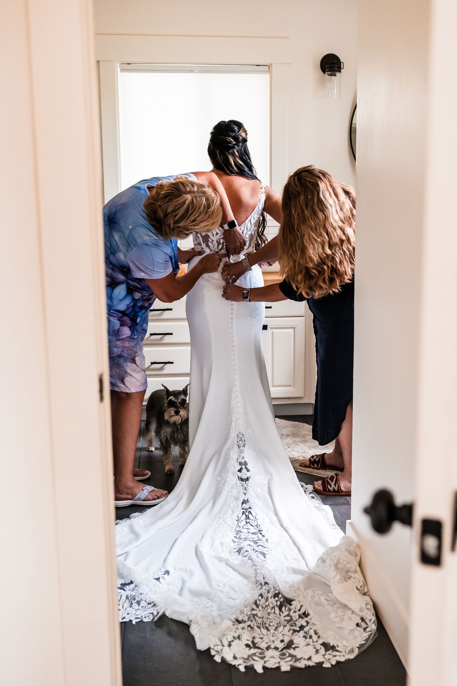 Two women help a bride zip up her lace wedding gown while a small dog watches nearby.