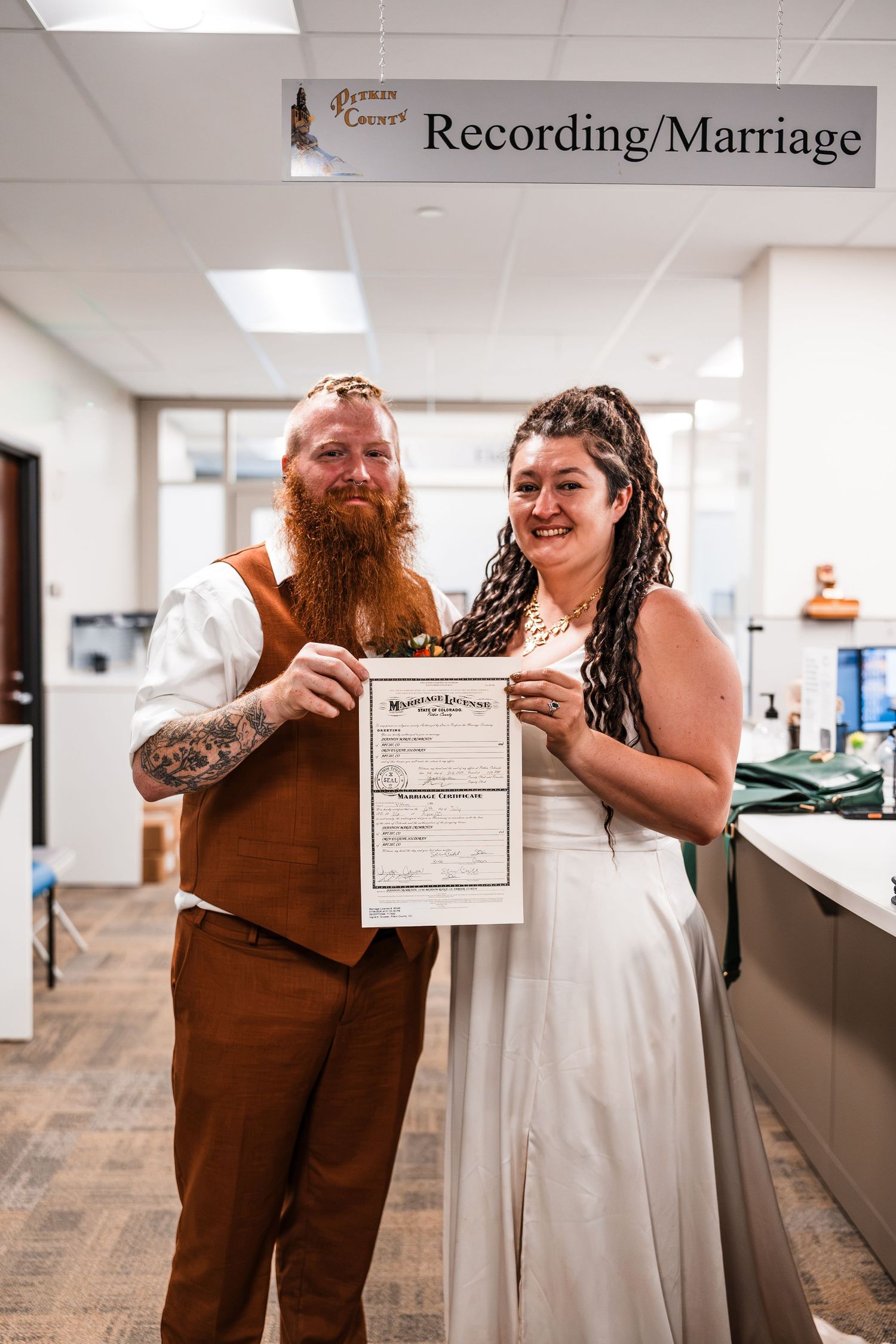 Smiling couple in wedding attire holds marriage certificate at Pitkin County Recording office counter.