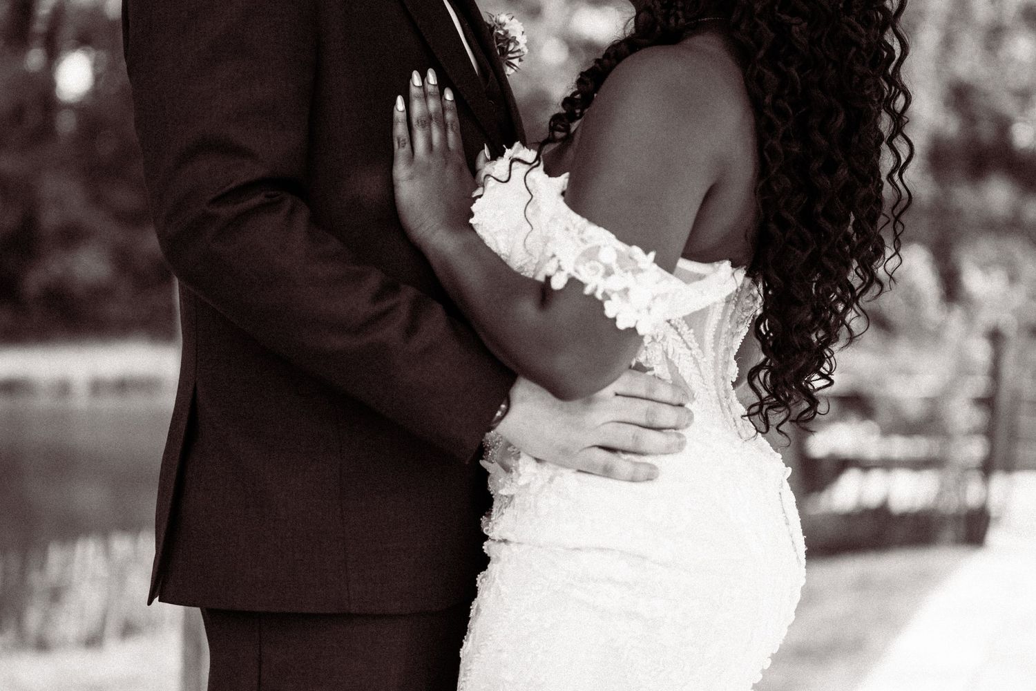 Black and white close-up of bride and groom embracing tenderly in wedding attire by a lakeside.